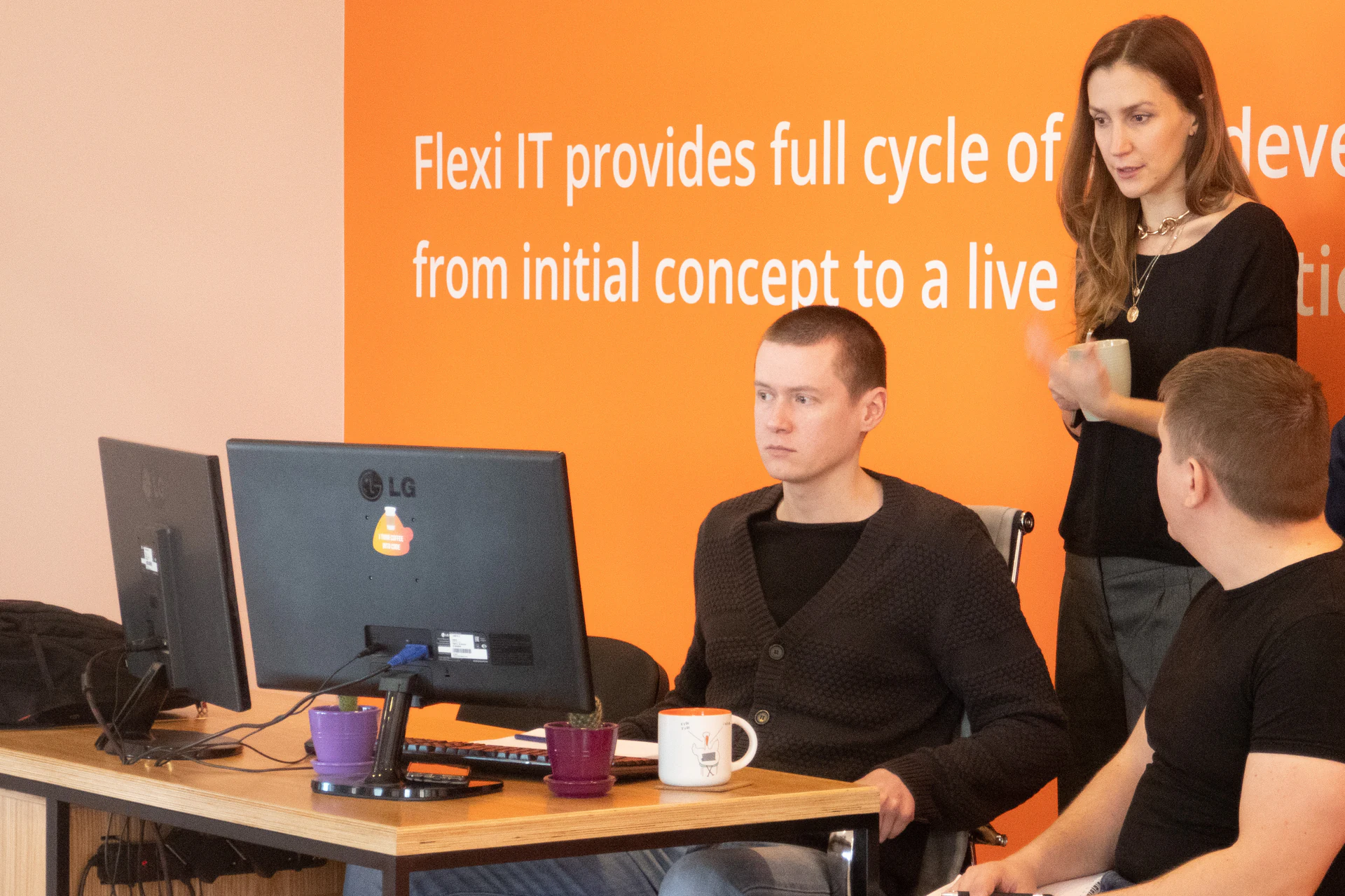 Flexi IT developers at a monitor in the office, a colleague with a mug standing behind them